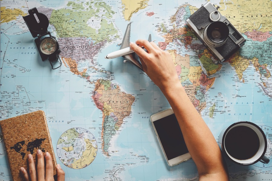 Grafika przedstawia przygotowania do podróży: mapa, notes w ręce, samolot w drugiej dłoni, a obok kompas i kawa.